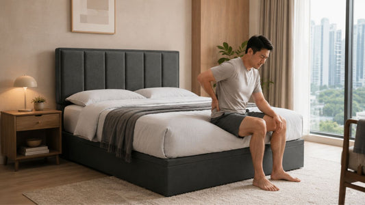 Man sitting on a grey upholstered bed in a modern Singapore bedroom while holding his lower back after waking up sore