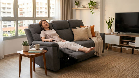 Woman relaxing on a grey massage recliner sofa in a warm Singapore living room with coffee table, shelving, and soft natural light